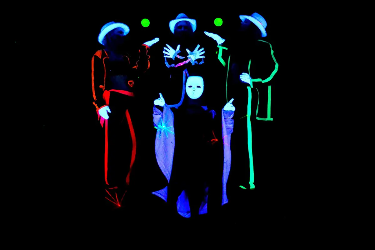 Fire optrædende i hatte og masker, der er omridset i glødende rødt, blåt og grønt, står mod en sort baggrund i et pragisk black light-show