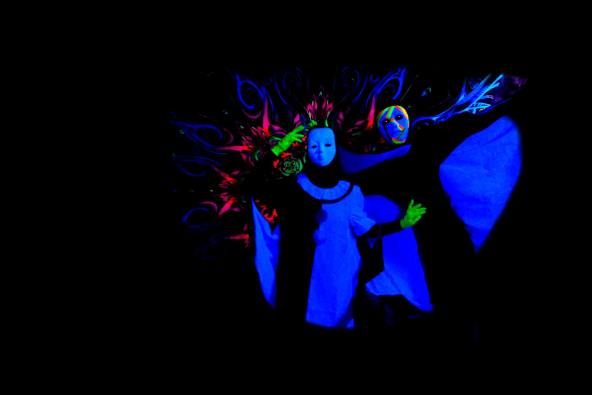 To maskerede performere i lysende blå kostumer og grønne handsker står foran en farverig fluorescerende baggrund i et mørkt black light-teater i Prag